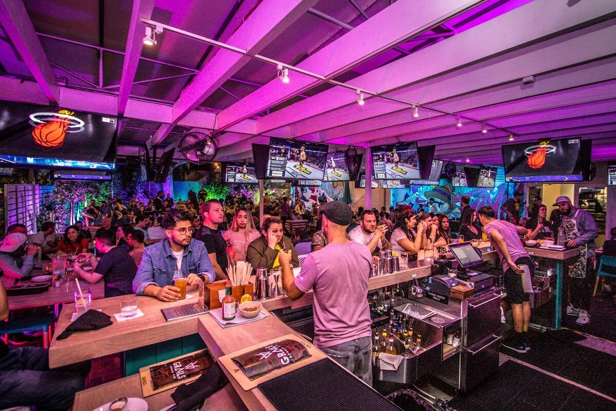 Crowded sports bar with neon pink ceiling lighting, multiple TV screens, and patrons at bar counter during NBA game night.