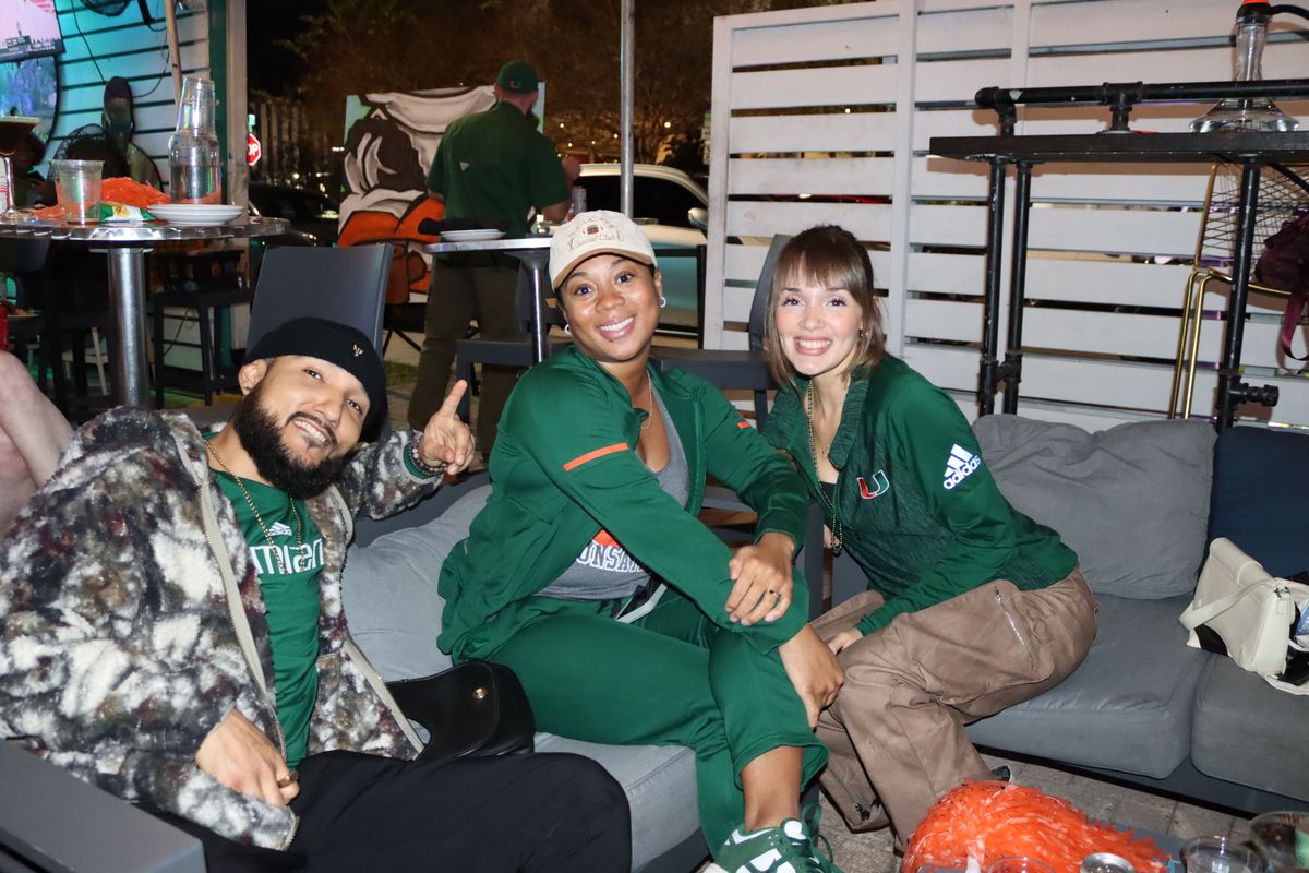 Three Miami Hurricanes fans in green and orange track suits relaxing on a Grails Miami patio couch on a Hurricanes game night