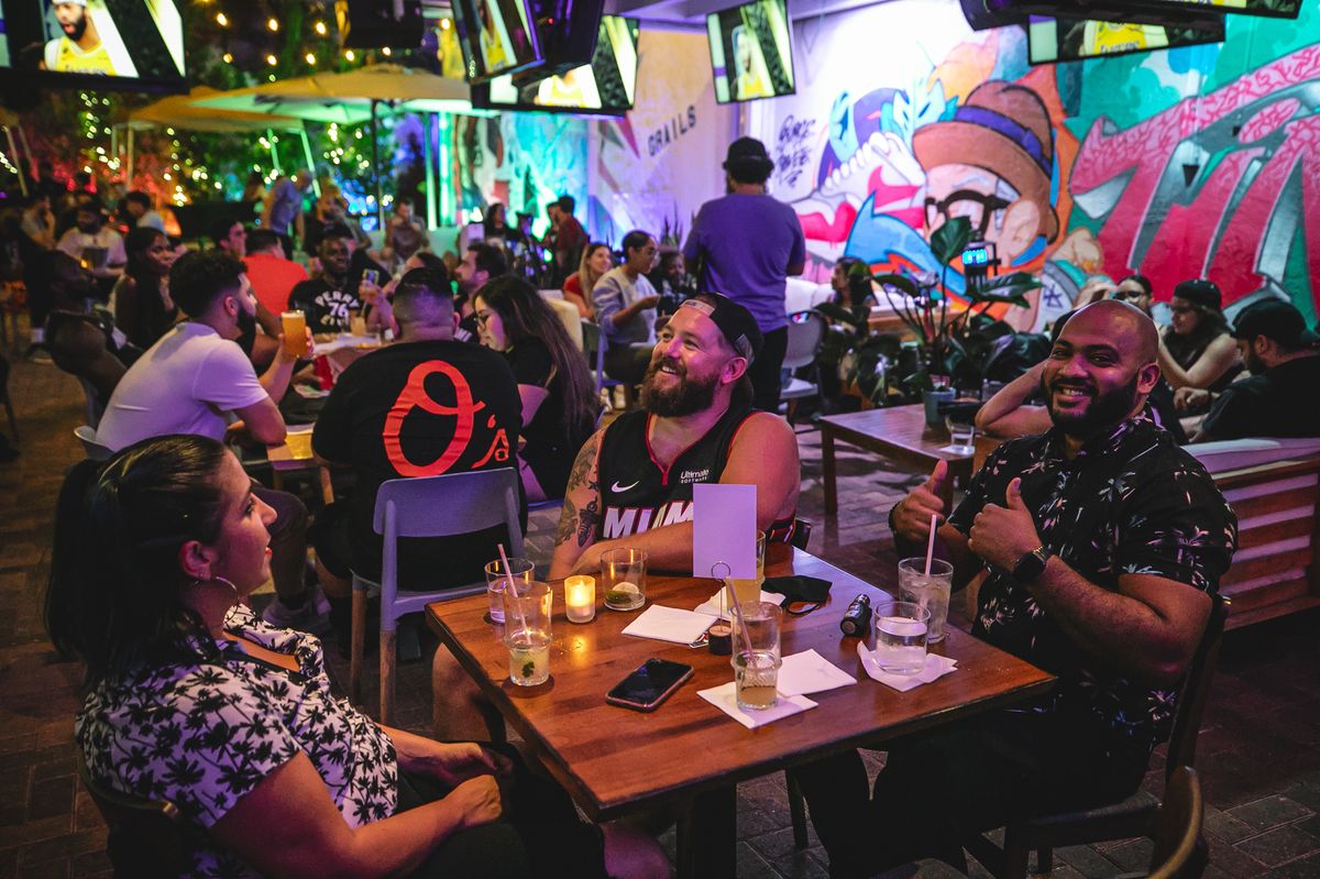 Three friends laughing at a Grails Miami patio table during an NBA night, one in a Miami Heat jersey