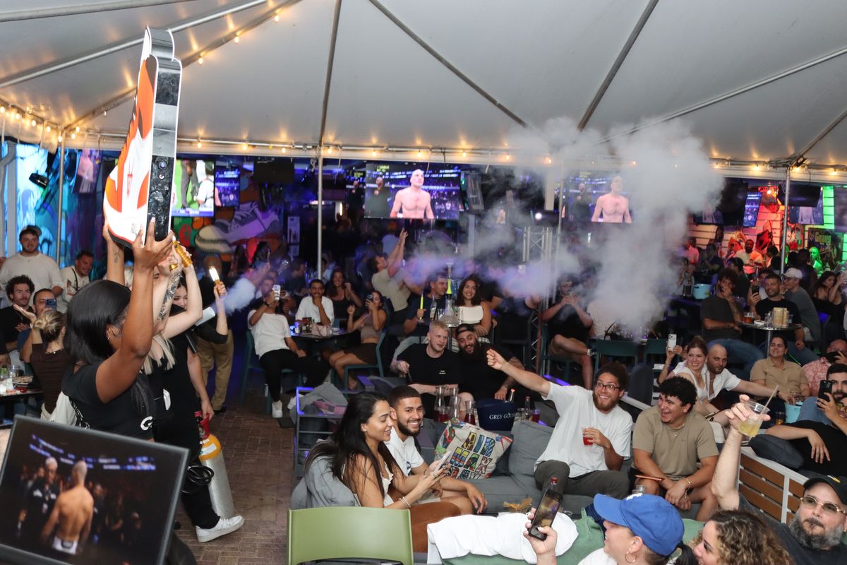 Crowd cheering at a UFC watch party as servers present sneaker cocktails on the Grails patio.