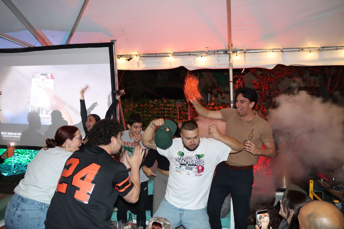 Miami Hurricanes fans cheering with orange pom-poms and a Hurricanes cap at Grails Miami during a College Football Playoff watch party under the tented patio