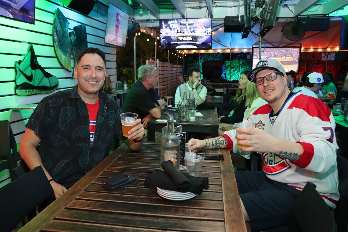 Two friends at a Grails Miami table with NHL playoff hockey on overhead TVs, one wearing a Montreal Canadiens jersey and cap during a Stanley Cup Playoff watch party