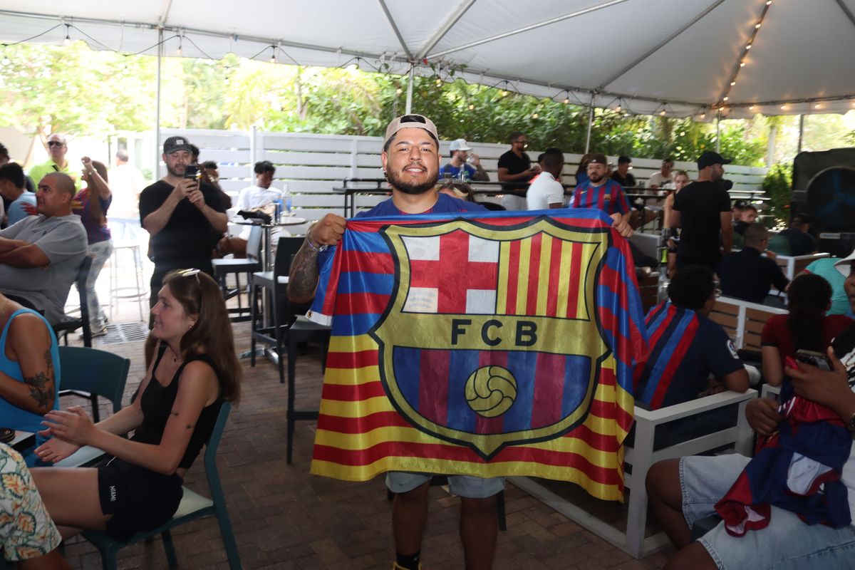 Man in an FC Barcelona jersey holding up a large FCB crest flag during a Champions League watch party at Grails Miami