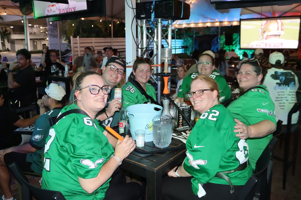 Philadelphia Eagles fans in green team jerseys smiling around a patio hookah table while the NFL game plays on TVs behind them at Grails Miami