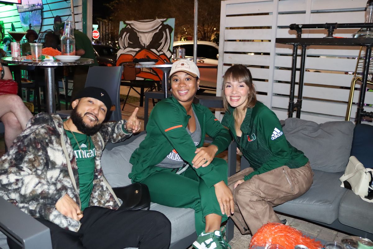 Three Miami Hurricanes fans in green UM gear posing together on a patio couch at Grails Miami in front of the orange-and-green Ibis mascot mural during a college football Saturday