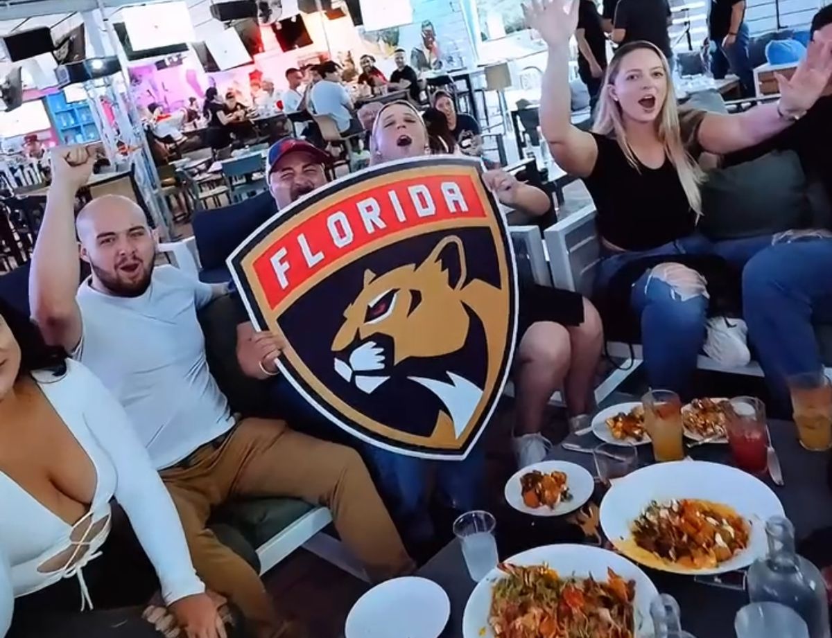 Group of Florida Panthers fans cheering around a table with food, cocktails and a Panthers logo sign at Grails Miami during a Stanley Cup Playoff watch party