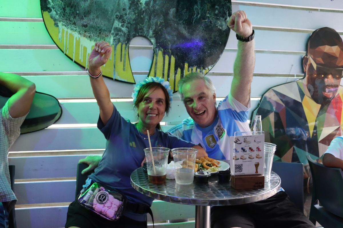 Couple in soccer jerseys cheering with raised fists at their table during a watch party at Grails Miami, the sports bar near Edgewater