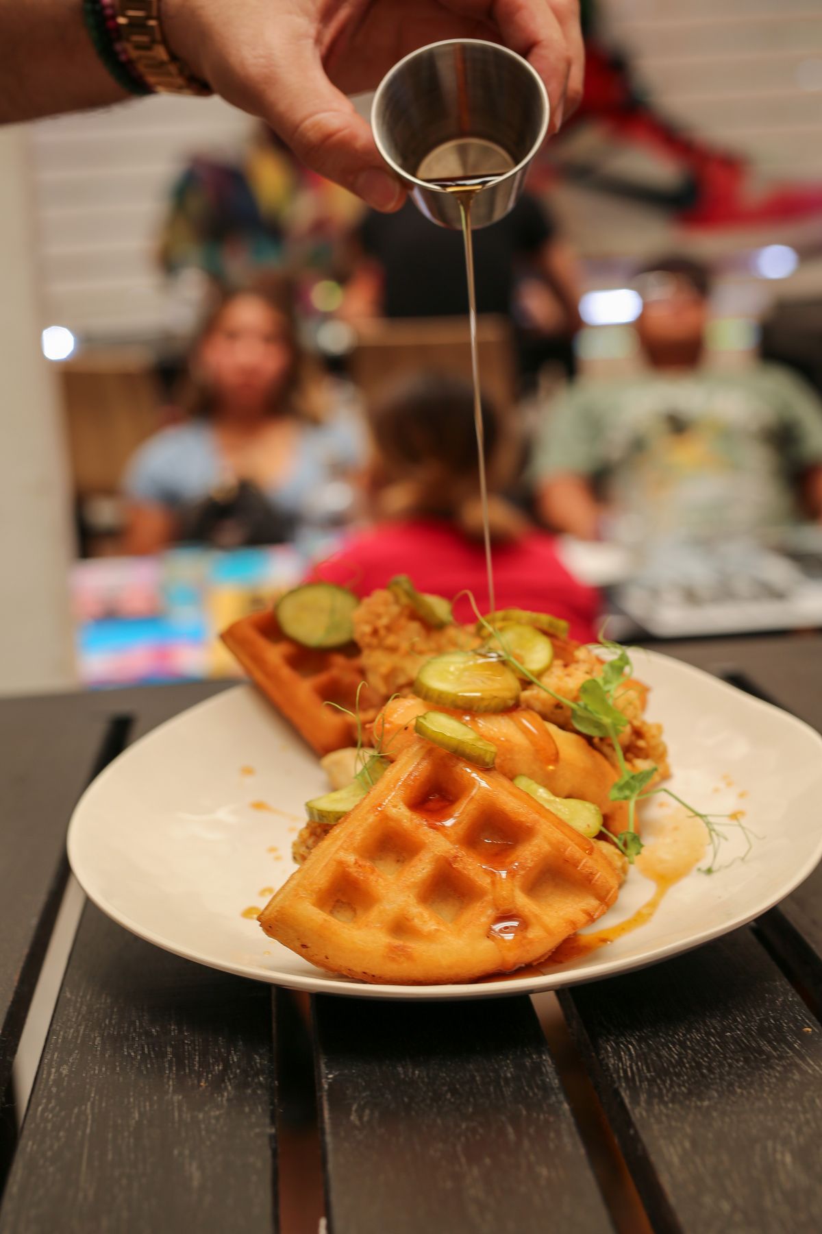 Hand pouring syrup from a jigger onto chicken and waffles at Grails Miami Sunday brunch.