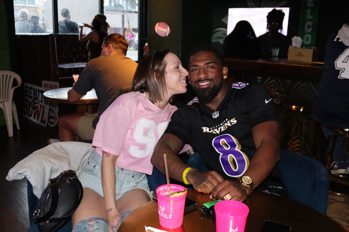 Couple in Baltimore Ravens jerseys posing at a Grails Miami table with pink Grails cocktail cups during an NFL watch party