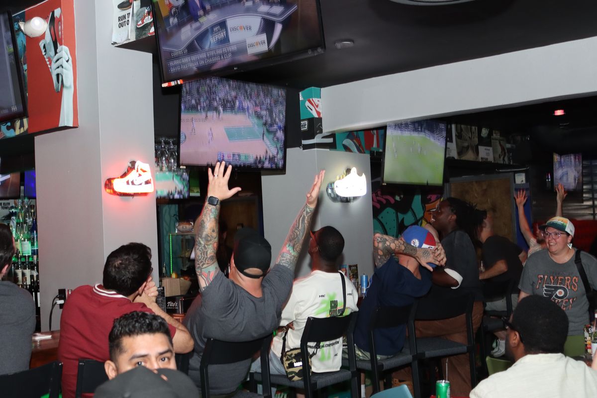 Fans with arms raised cheering at a basketball broadcast on the TVs above the bar at Grails Miami, Miami's home WNBA bar