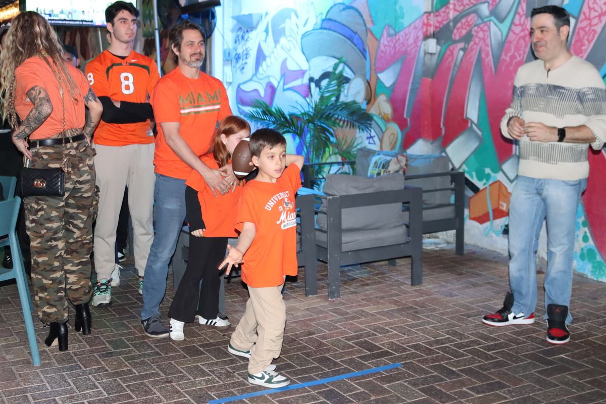 Kid in an orange Miami Hurricanes shirt throwing a football inside Grails Miami while a family of Hurricanes fans cheer