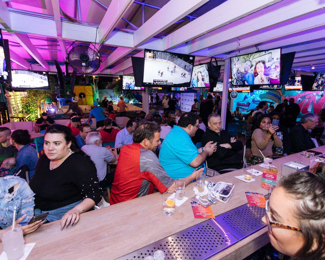 Crowd of fans seated along the bar at Grails Miami watching an NHL hockey game on overhead TVs during a Stanley Cup Playoff watch party