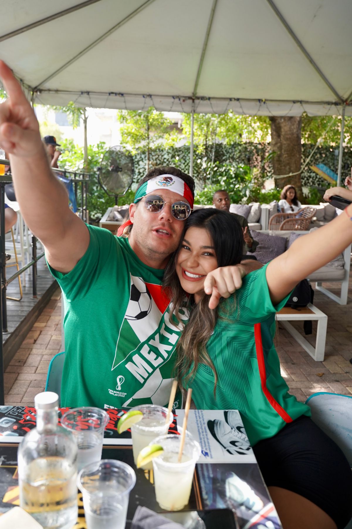 Couple in Mexico jerseys posing with arms raised at the Grails Miami patio during a Concacaf Champions Cup Liga MX-club watch party