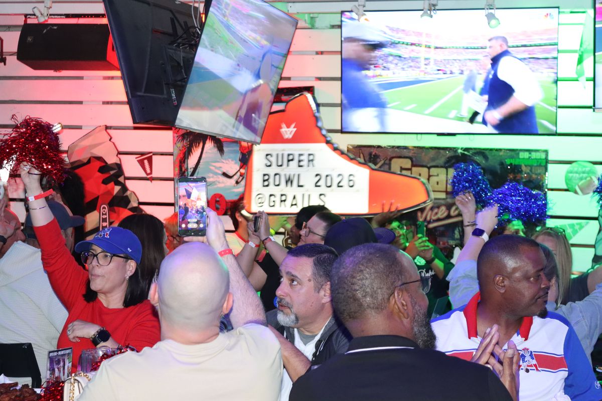 New England Patriots fans cheering with pom-poms beneath the glowing sneaker sign at Grails Miami during a Super Bowl watch party