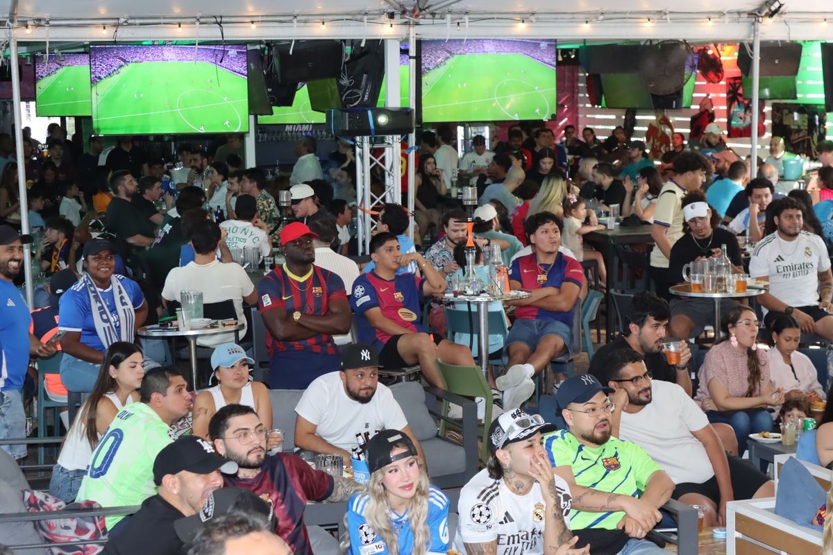 Wide view of a crowd of fans in Real Madrid and FC Barcelona jerseys watching El Clasico on dual large screens under the tented patio at Grails Miami Wynwood during a Champions League soccer watch party.