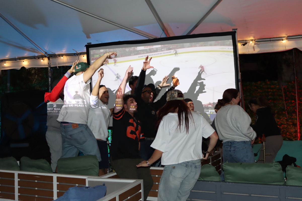 Hockey fans cheering with arms raised in front of a giant projection screen during a Stanley Cup Playoff watch party at Grails Miami