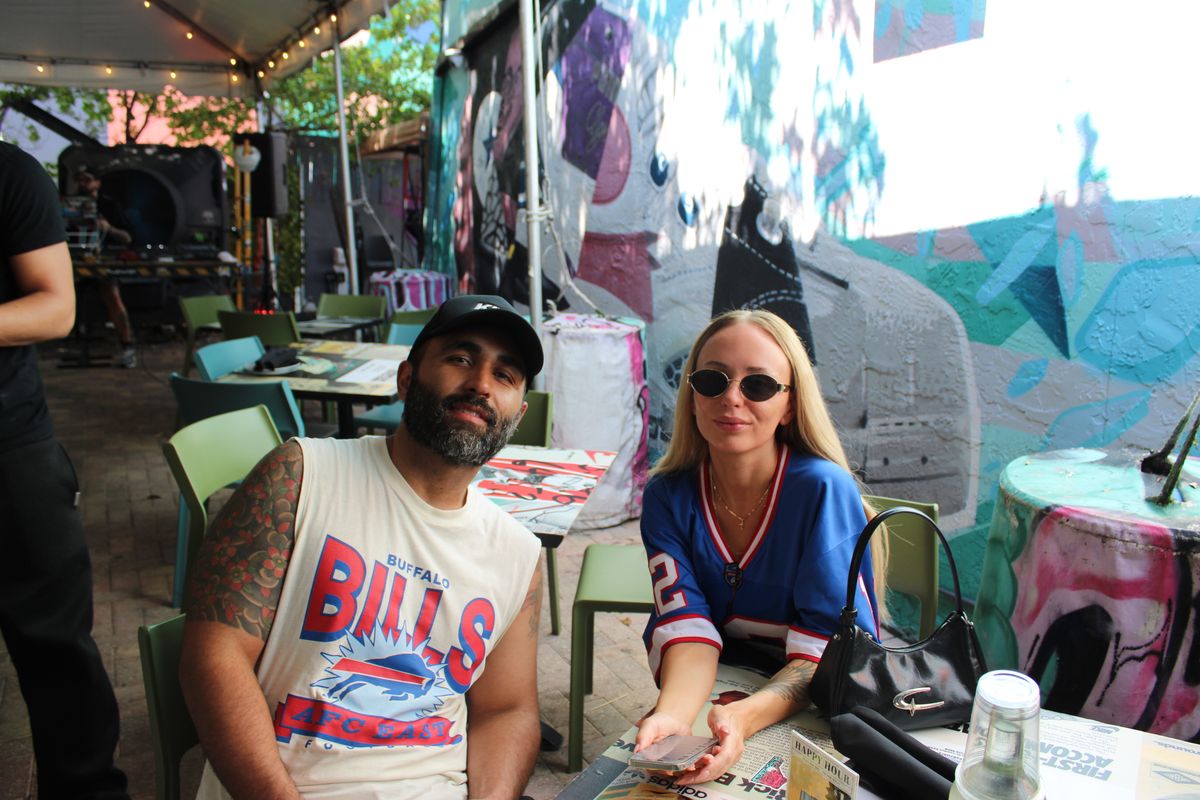 Couple in a Buffalo Bills tee and a Bills jersey at a covered patio table under the Grails Miami mural on an NFL Sunday