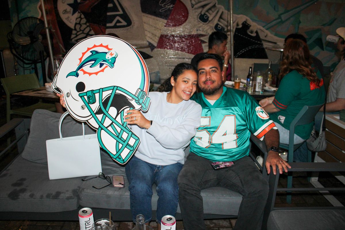 Miami Dolphins fan couple posing with an oversized Miami Dolphins helmet cutout at Grails Miami, the husband wearing a teal #54 Dolphins jersey