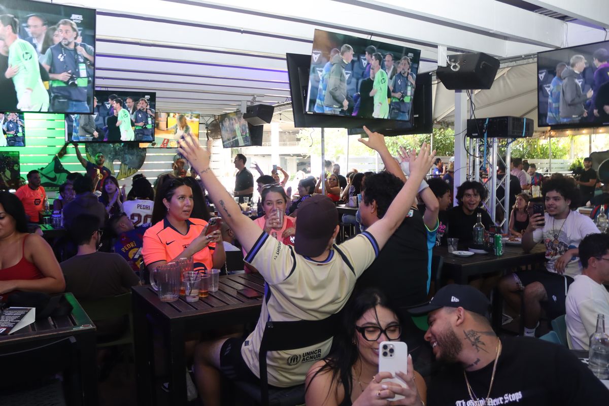 Football fan in a cream jersey raising both arms in celebration at a packed Serie A watch party at Grails Miami