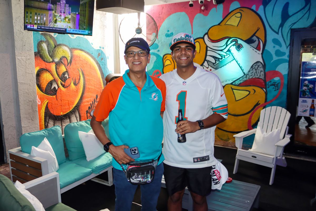Father and son in Miami Dolphins jerseys posing next to a Grails Miami mural before kickoff
