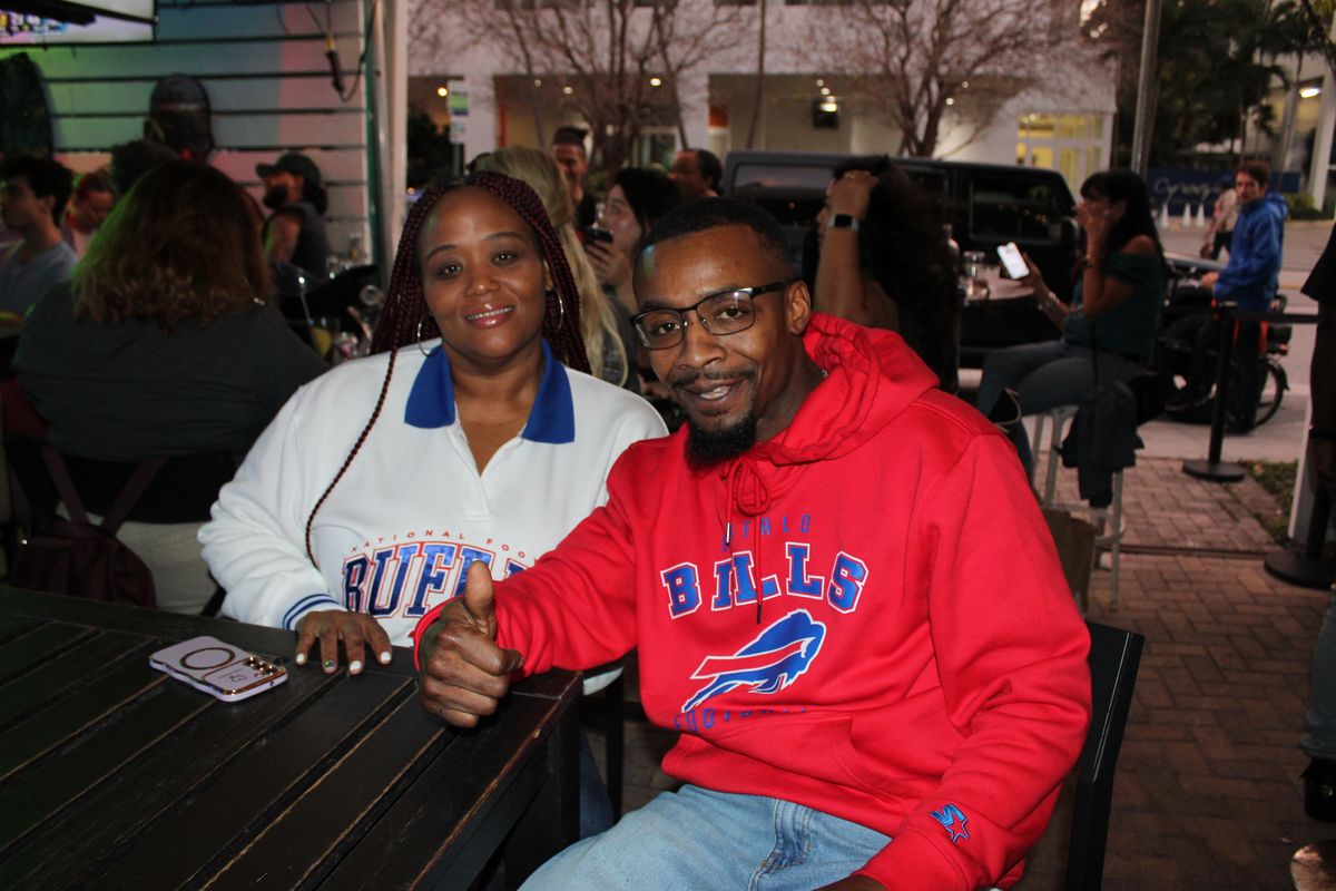 Couple in a red Buffalo Bills hoodie and a Buffalo varsity jacket at a Grails Miami patio table on a Bills game night in Wynwood