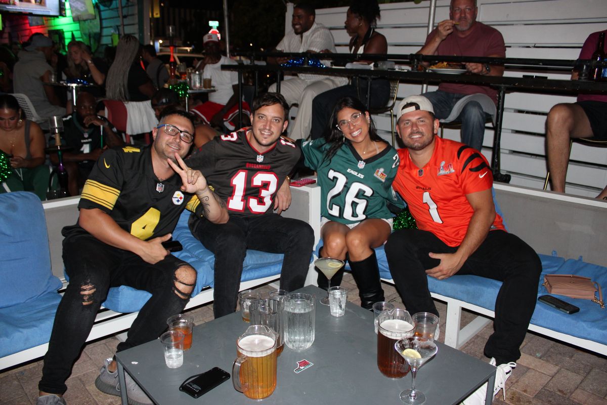 Four NFL fans in Steelers, Buccaneers, Eagles and Bengals jerseys posing together with beers and martinis at Grails Miami during an NFL watch party