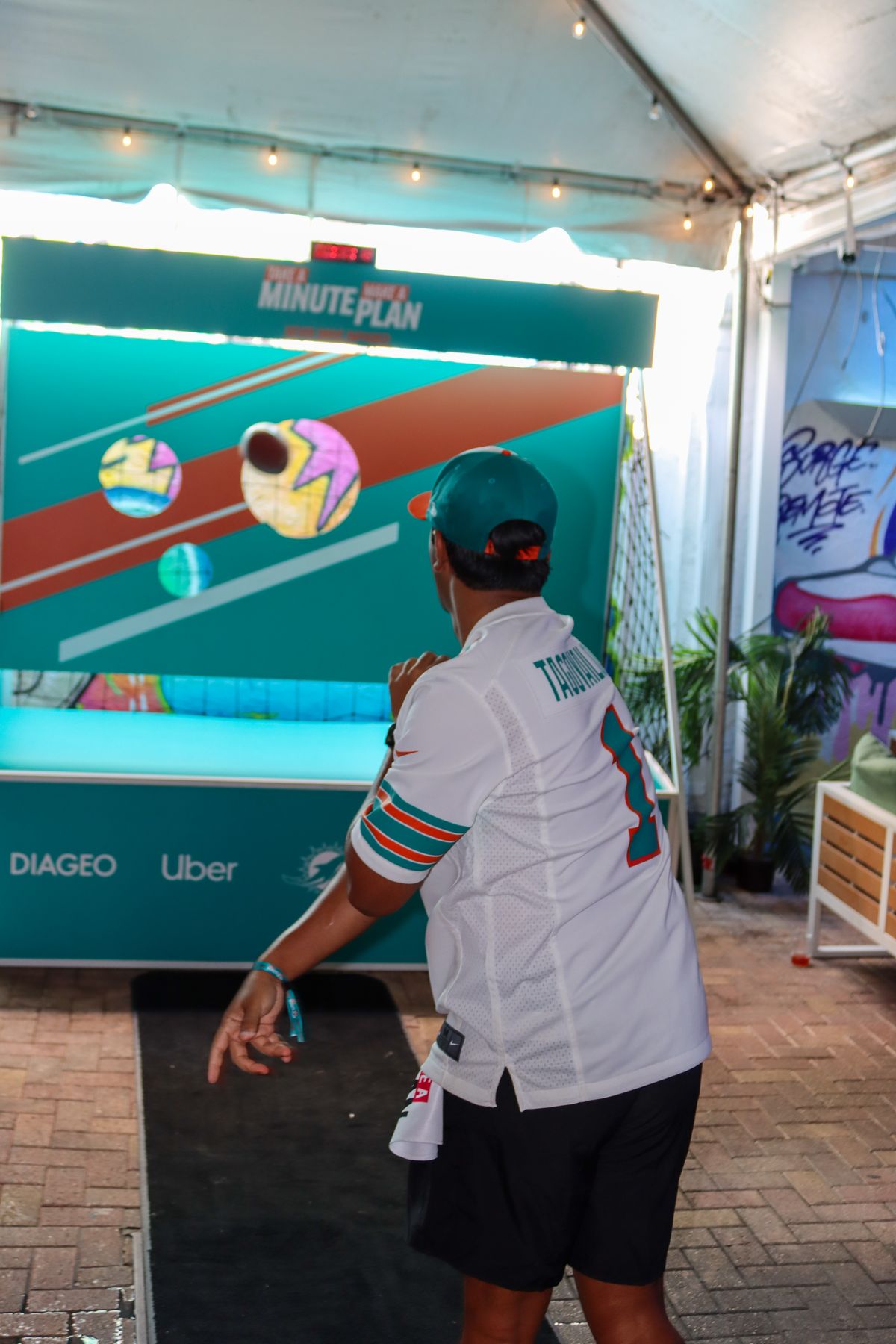 Fan in a Tua Tagovailoa Miami Dolphins jersey throwing a mini football at an interactive Take A Minute Make A Plan target game at Grails Miami