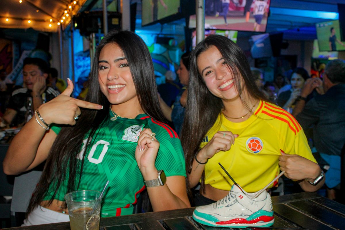 Two women in Mexico and Colombia soccer jerseys posing at a Grails Miami table during a Concacaf Gold Cup watch party
