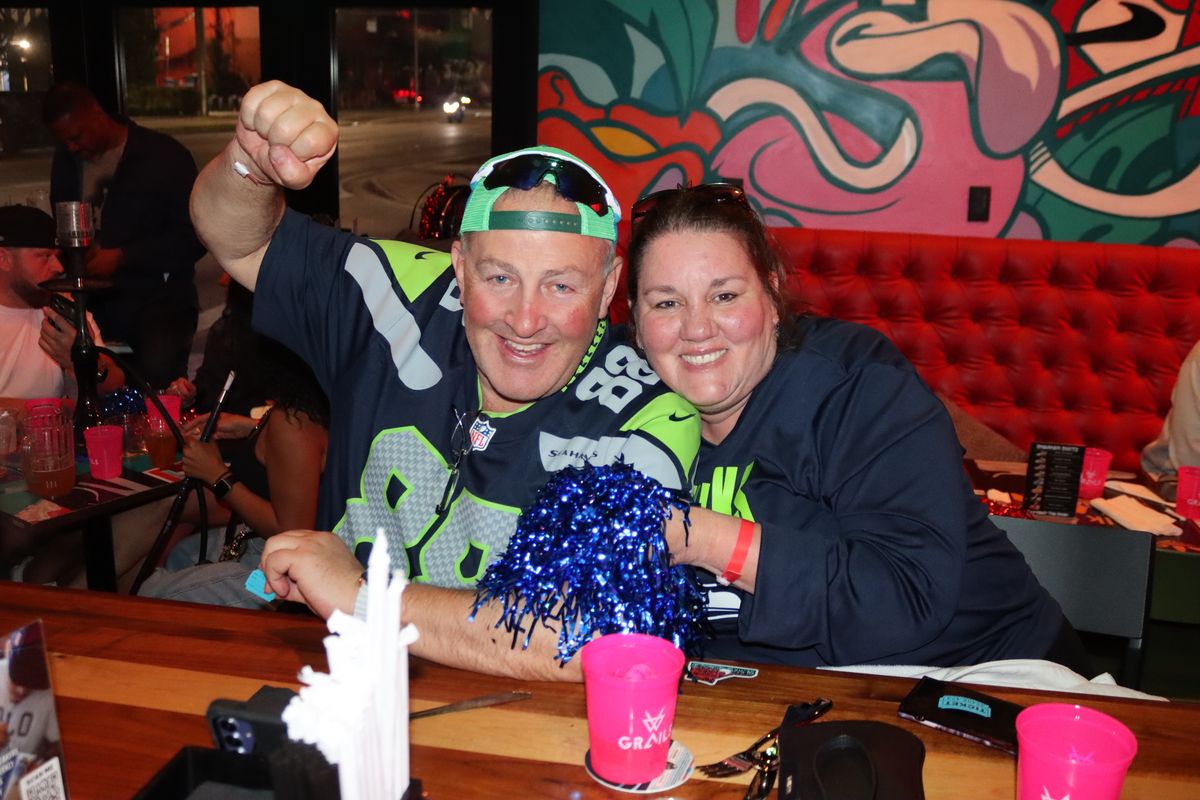 Seattle Seahawks fan couple in team jerseys celebrating with a pom-pom and a raised fist at a Grails Miami booth during an NFL Sunday