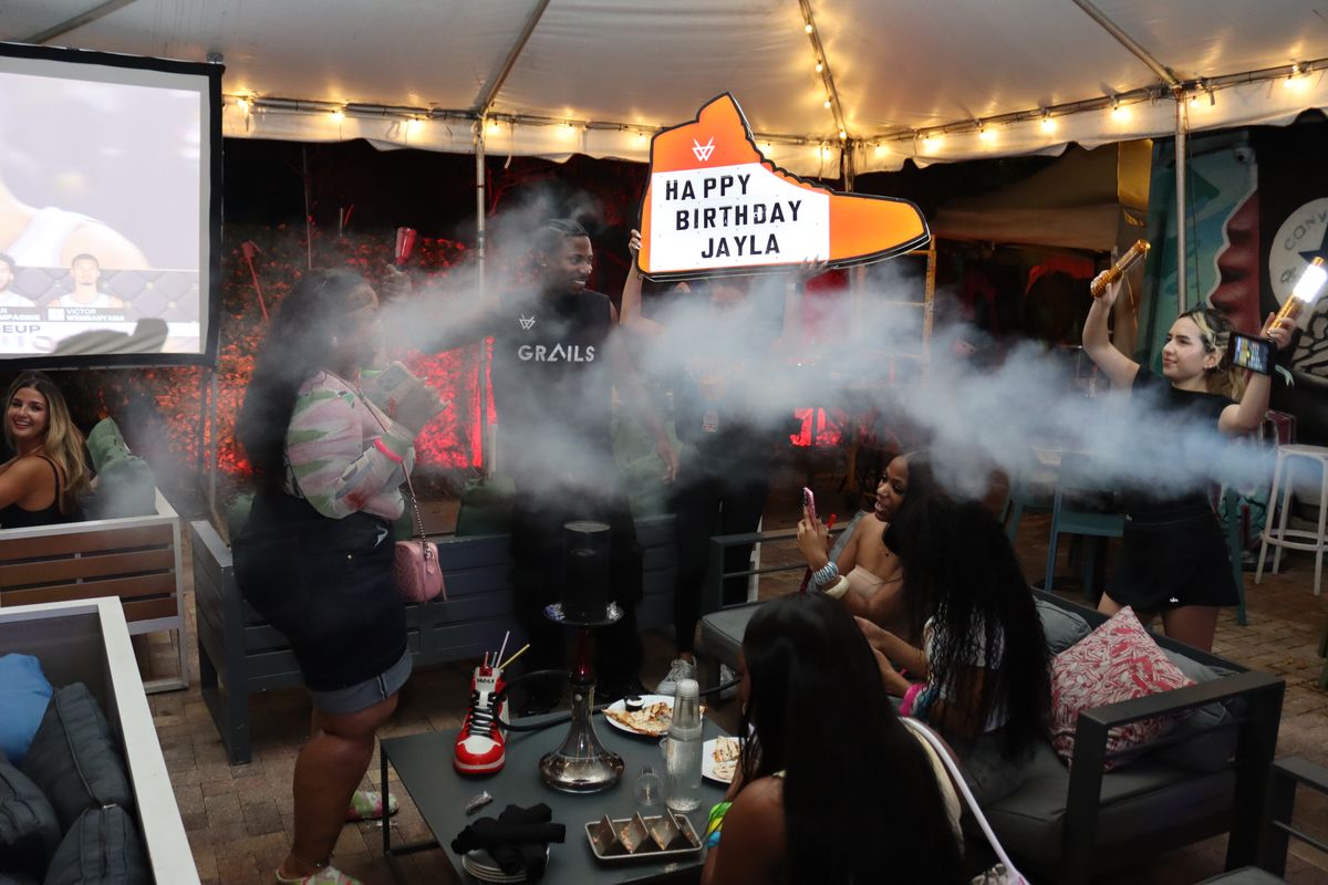Birthday group on the Grails Miami patio with hookah smoke and an illuminated sneaker sign — birthday hookah party in Miami