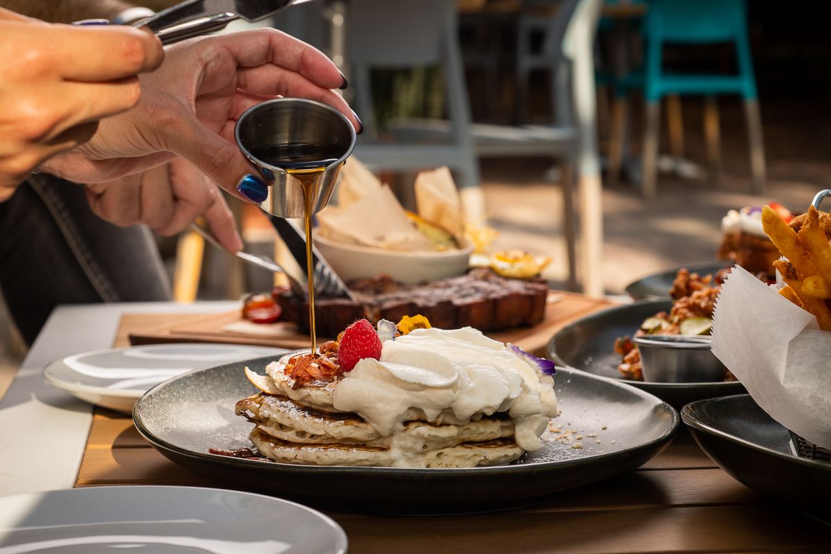 Hand pouring syrup onto a stack of pancakes topped with whipped cream and berries at Grails Miami Sunday brunch.