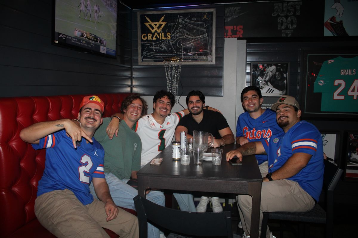 Six friends in Florida Gators and Miami Hurricanes jerseys sharing a red booth under the Grails Miami sign with college football on the TV