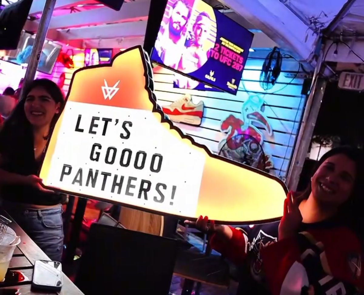 Two Florida Panthers fans holding a glowing sneaker-shaped LET'S GO PANTHERS sign at Grails Miami during a Florida Panthers watch party