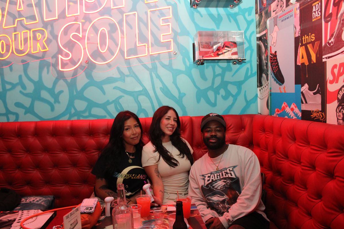 Three friends, one in a Philadelphia Eagles sweatshirt, in a red booth under the neon sign in the indoor sneaker lounge at Grails Miami