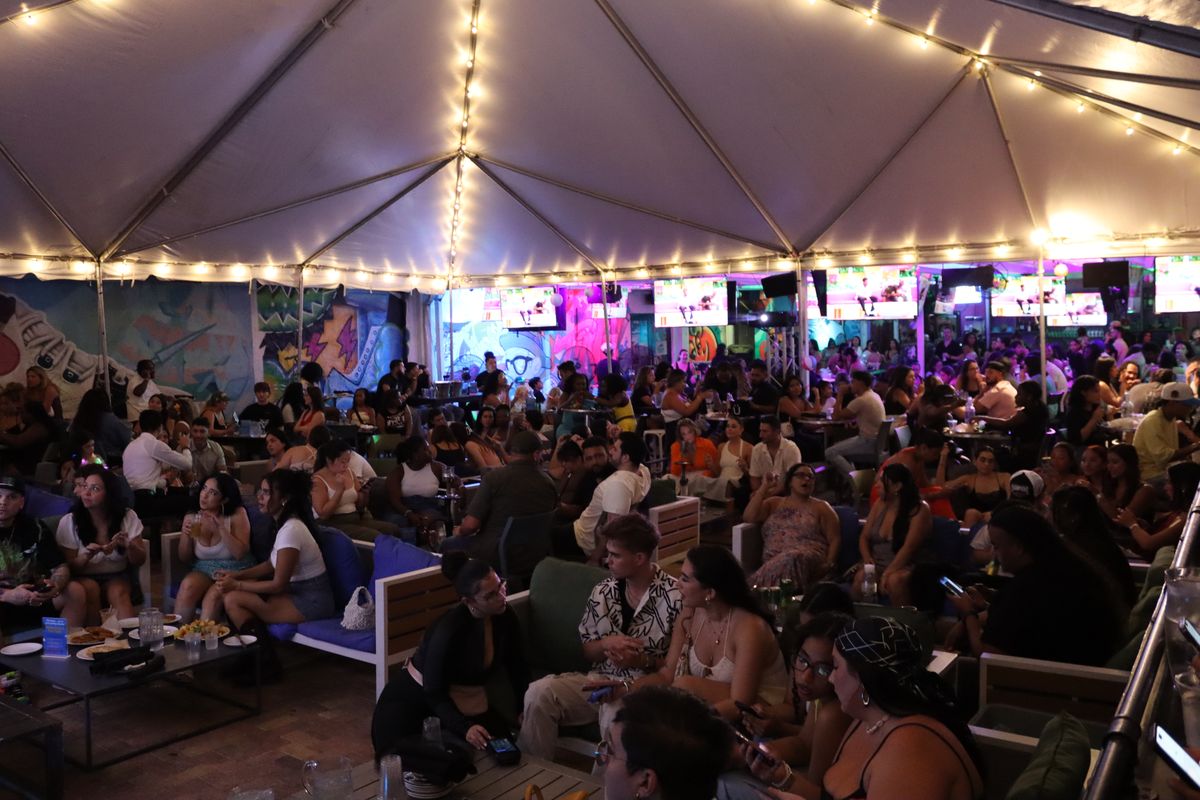 The Grails Miami covered Miami Vice patio at night: a packed crowd on lounge couches under a tented, string-lit roof with multiple TVs and Wynwood murals