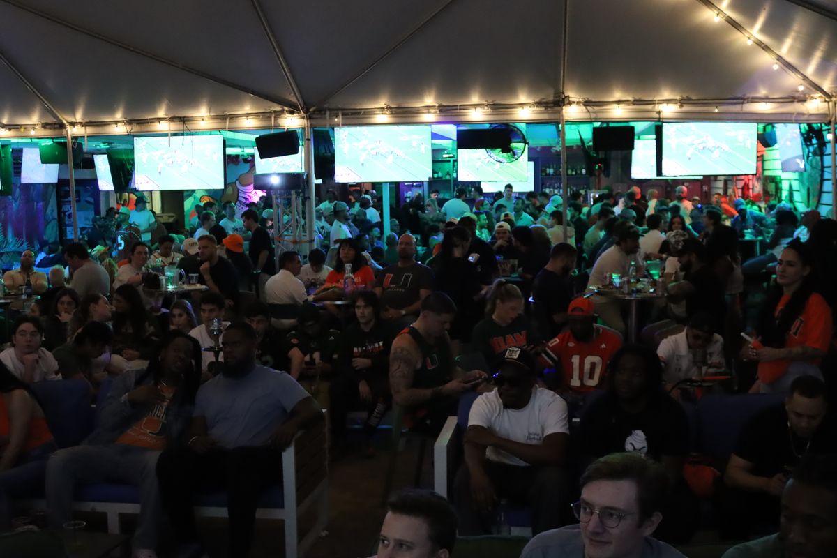 College football fans in Miami Hurricanes and other NCAA jerseys watching the Playoff under the giant Grails Miami tent