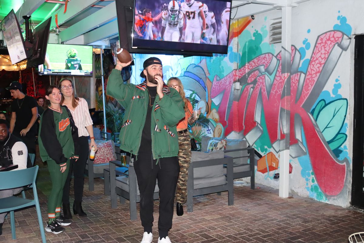 Miami Hurricanes fan in a green Hurricanes jacket holding a football while a Miami Hurricanes game plays on a Grails Miami TV