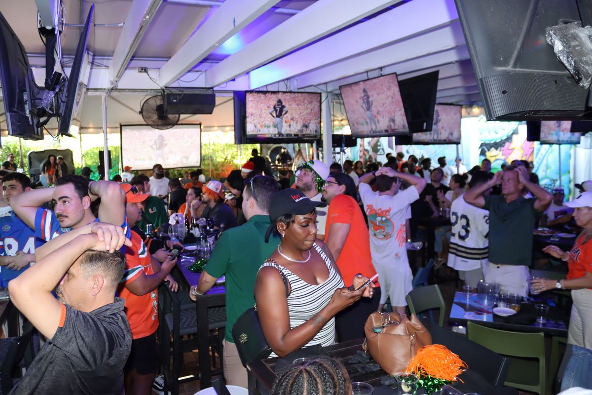 Mixed crowd of Miami Hurricanes and rival college football jerseys reacting to a Playoff play under the Grails Miami tent