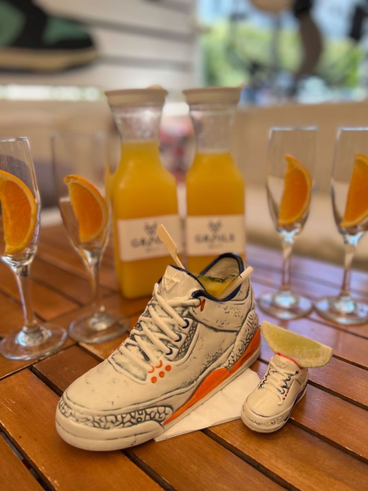 Sneaker-shaped ceramic cocktail beside Grails-branded mimosa bottles on a wooden patio table — the Wynwood brunch signature.