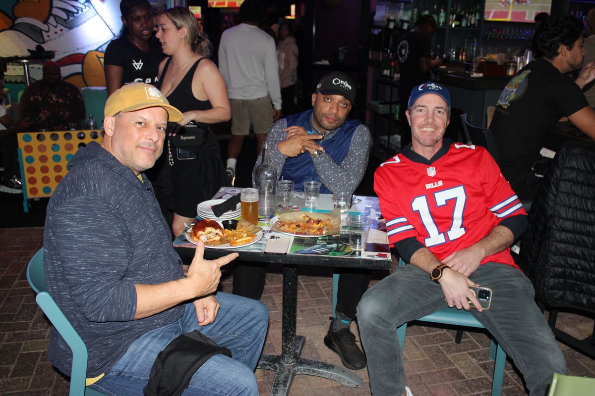 Three friends with nachos and beers at a table, one in a red Buffalo Bills number 17 jersey, watching NFL football at Grails Miami