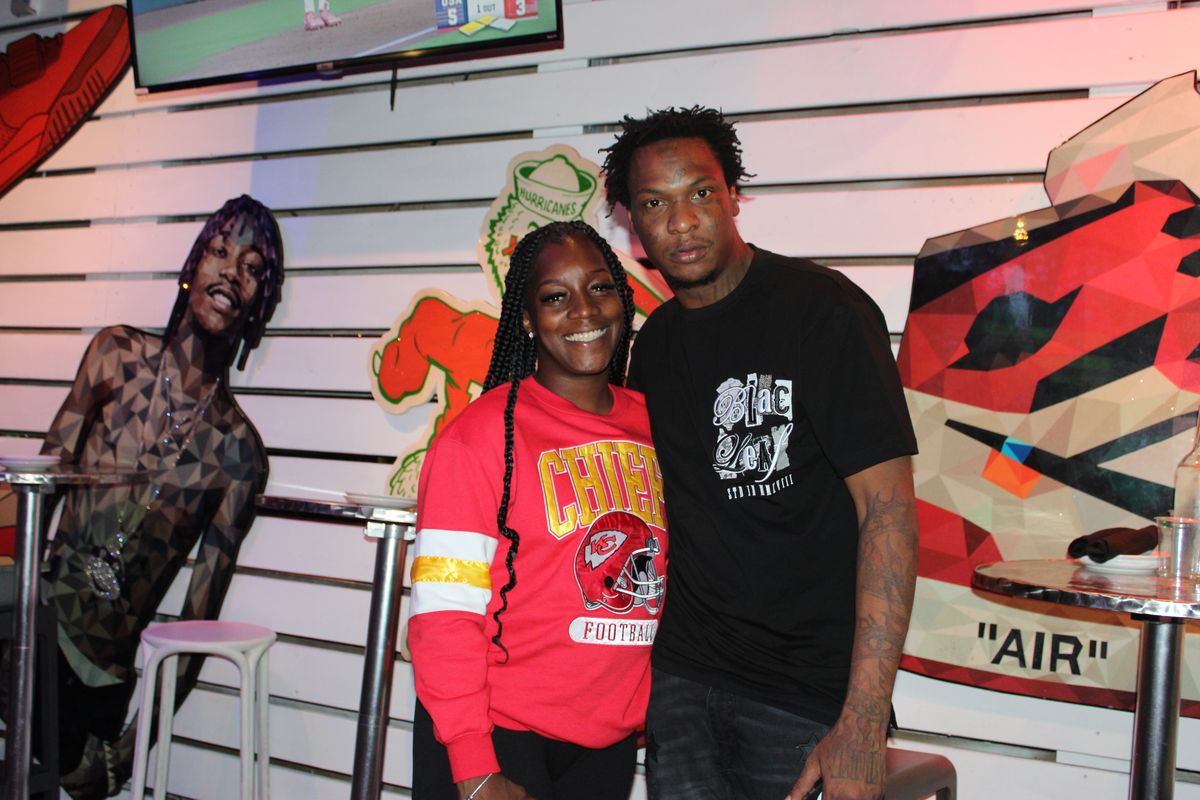 Couple in Kansas City Chiefs gear posing by the sneaker murals during an NFL Thursday night watch party at Grails Miami