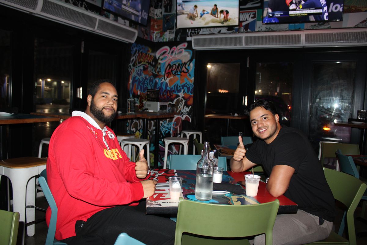 Two friends giving a thumbs up at their table in the indoor sneaker lounge, one in Chiefs colors, at Miami's Monday Night Football bar