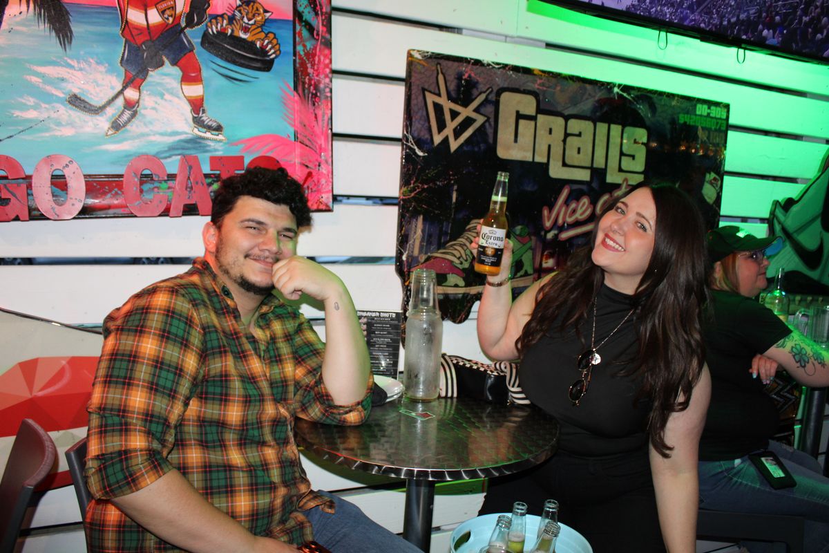 Smiling couple at a Grails Wynwood table with a Corona beer raised in front of a Florida Panthers mural