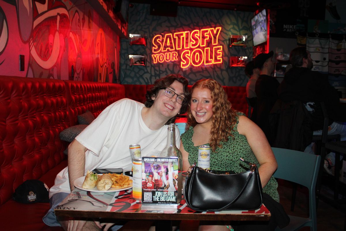 Couple posing at a red tufted booth at Grails Wynwood with cocktails and a plate of food under the Satisfy Your Sole neon sign on a World Series game night