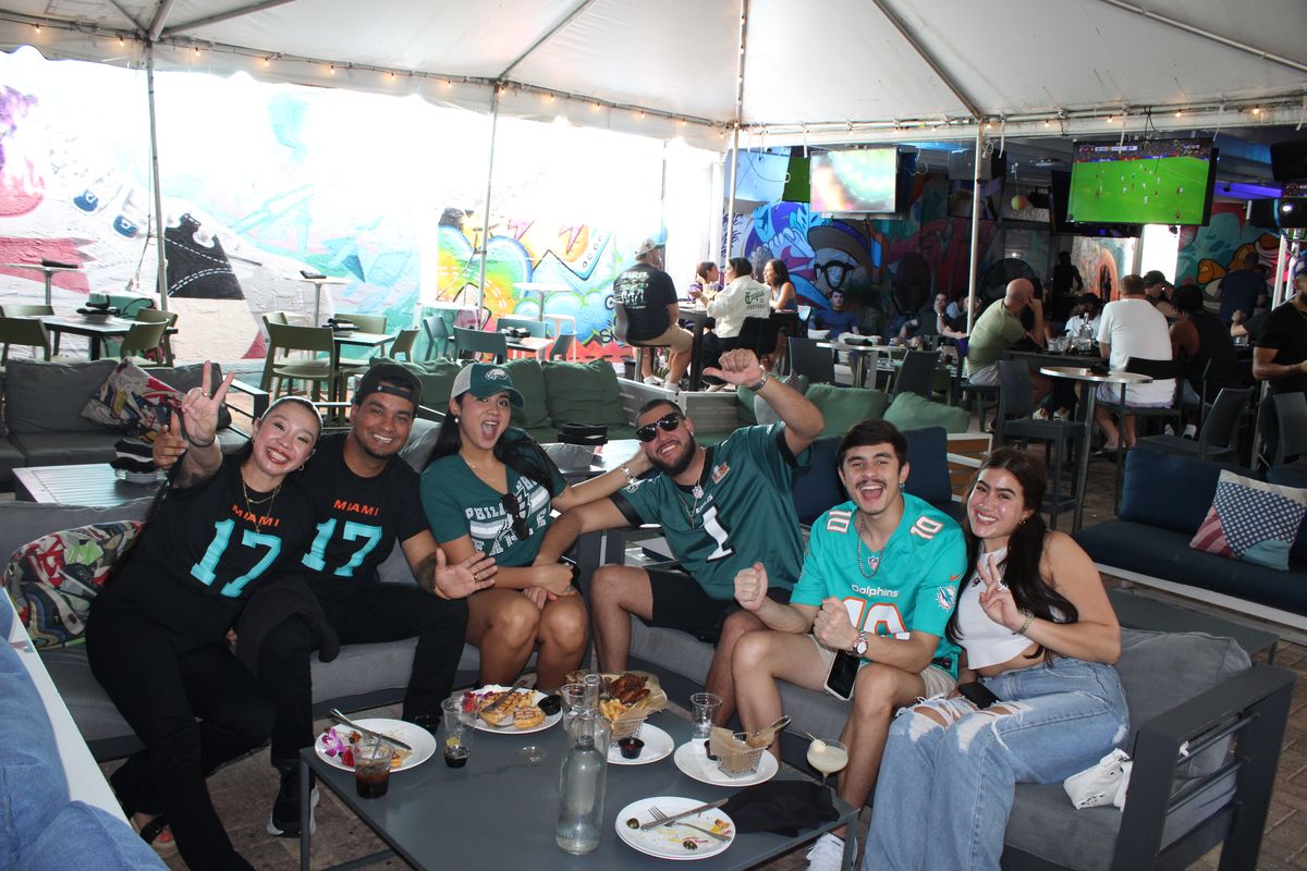 Six friends in Philadelphia Eagles and Miami Dolphins jerseys sharing a patio couch during an NFL watch party at Grails Miami