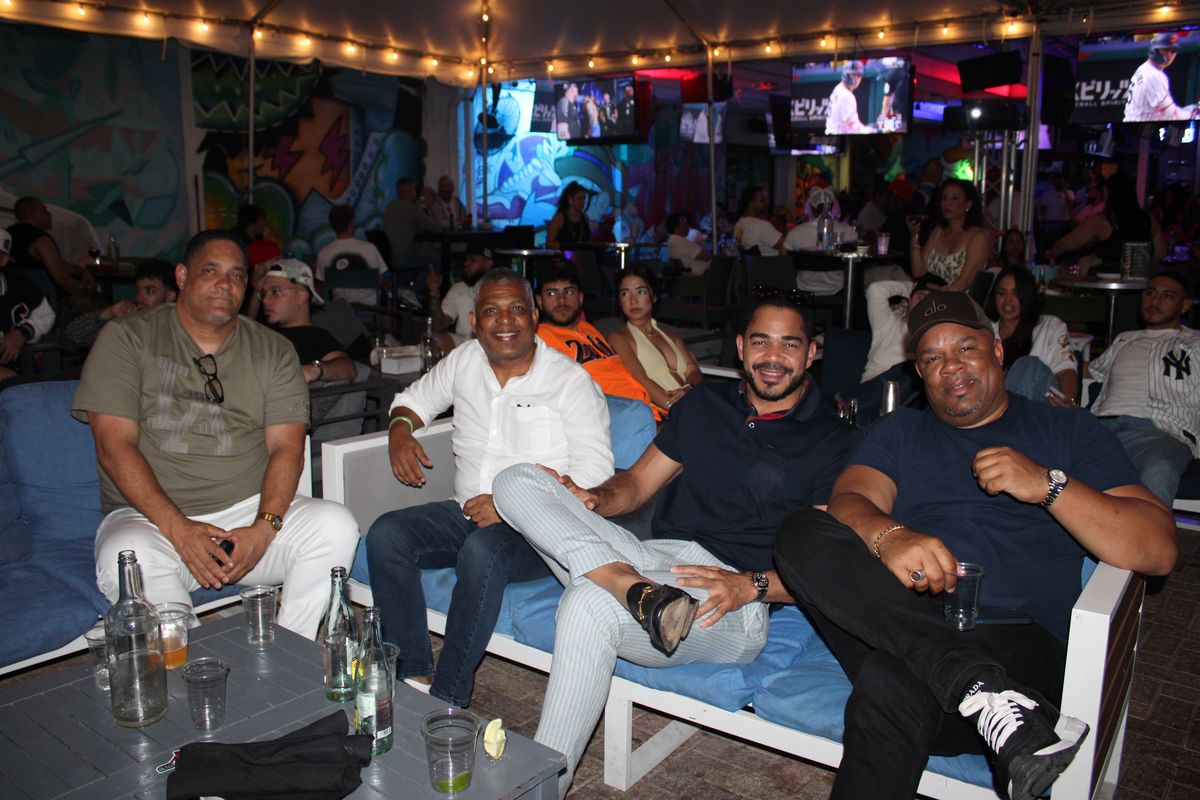 Four friends relaxing on a blue lounge couch at Grails Wynwood with bottles, glasses and baseball playing on overhead TVs during a World Series watch party