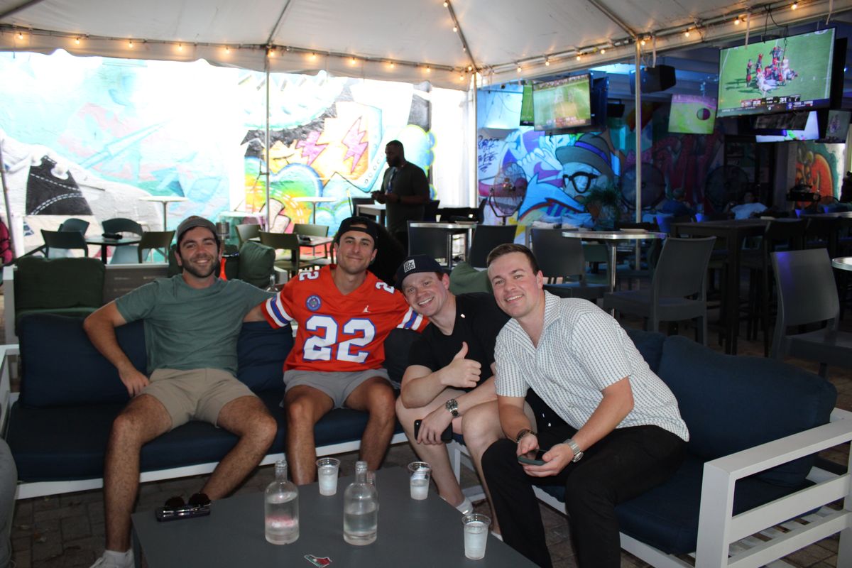 Four friends on the couches under the Grails Miami tent, one in a Florida Gators #22 jersey, with murals and TVs behind them