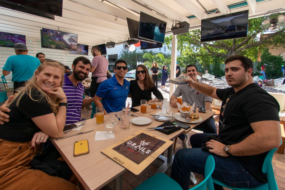 Group of six friends, one in a Florida Gators polo, around a patio table with beers and food at Grails Miami on a college football Saturday
