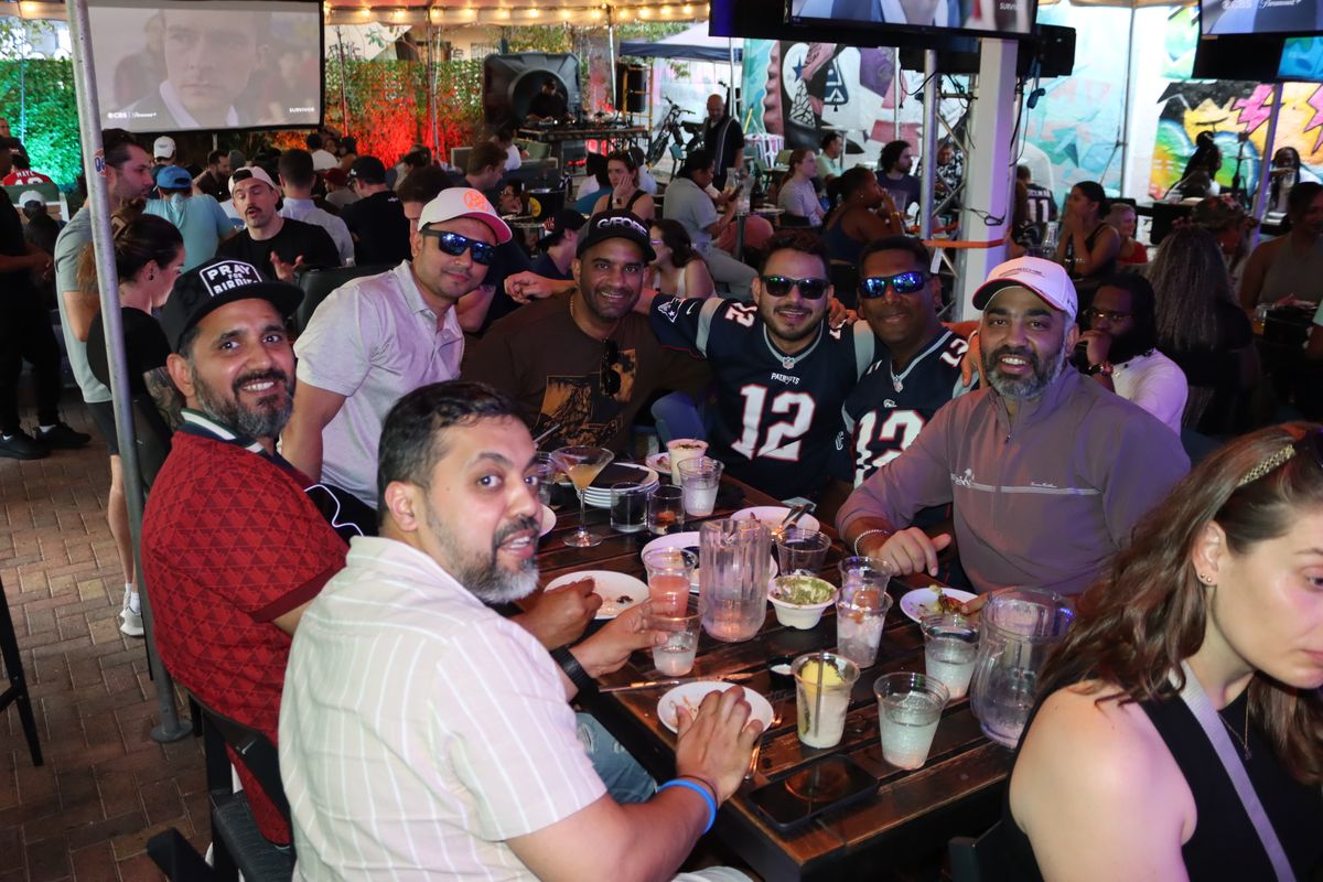 Group of friends, two in Patriots number 12 jerseys, sharing food and drinks at a long patio table during an NFL watch party at Grails Miami