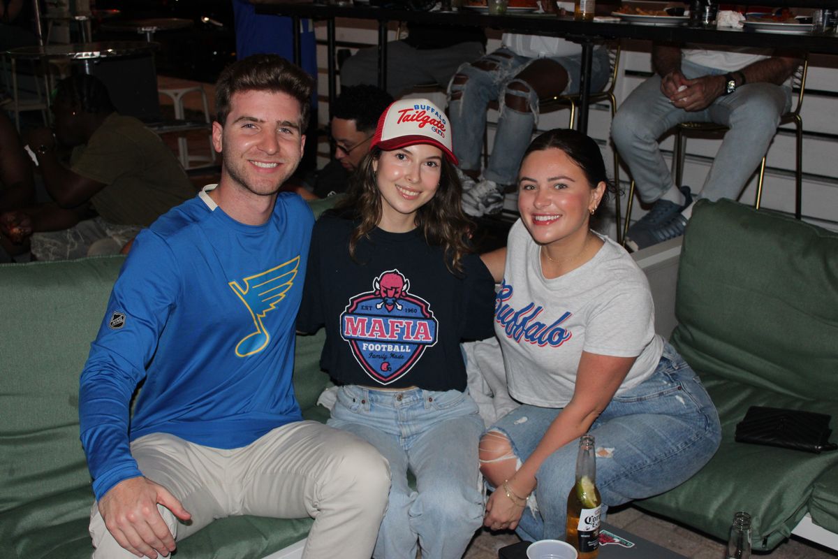 Three Buffalo Bills fans in Bills Mafia and Buffalo tees on a lounge couch at Grails Miami, the Buffalo Bills bar in Wynwood